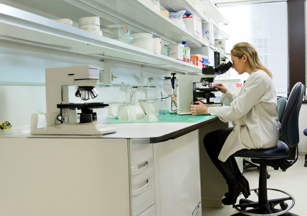 Female Scientist Working In Lab Looking Through Microscope