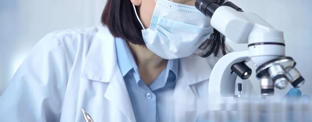 Female Scientist In Lab Goggles, Protective Mask And Gloves Is Using Microscope In Laboratory.